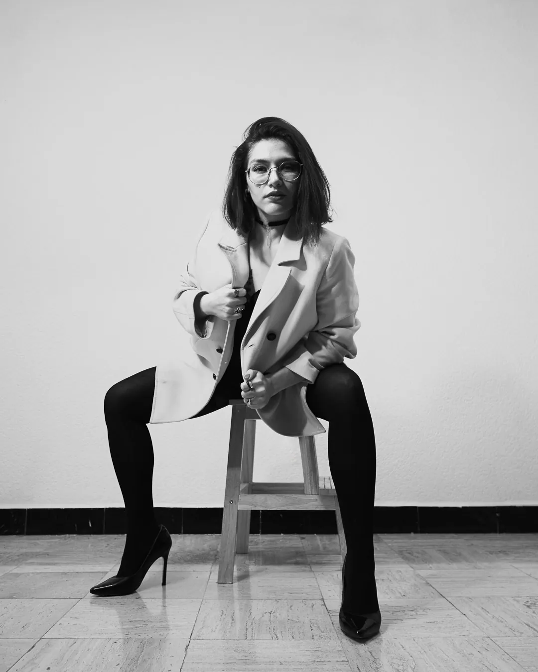 Mujer sentada en banco de madera con saco claro y medias negras, boudoir de estudio en blanco y negro