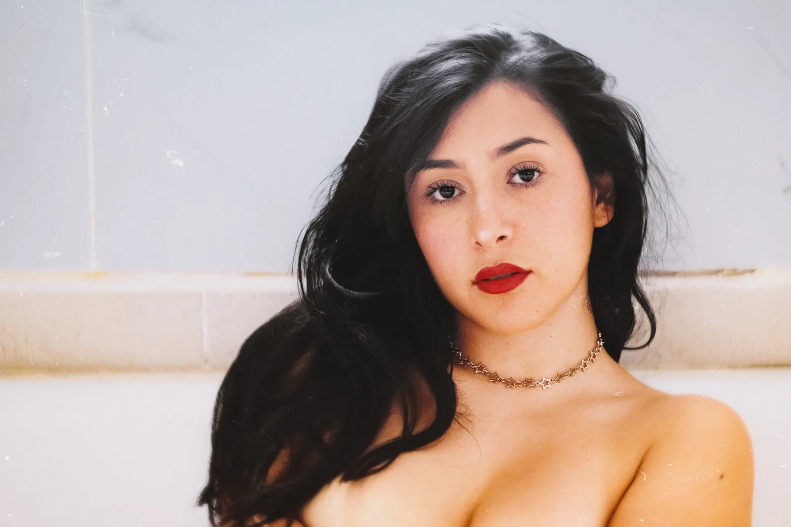 Retrato boudoir a color de mujer con cabello oscuro suelto, labios rojos y gargantilla de estrellas plateadas, recostada en una tina de baño durante una sesión de fotografía boudoir individual en Ciudad de México con Utopic Dystopia